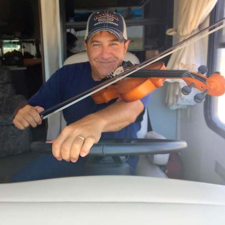 Musician Tim Cadiere smiles as he plays the fiddle at the wheel of a bus, pretending to drive.